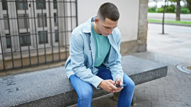 A young hispanic man in casual clothing is using a smartphone while sitting alone on a bench in an urban outdoor setting.