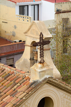 An ornate metal cross stands out against a backdrop of urban buildings; it’s perched atop an ancient roof with red tiles and detailed architectural structure.