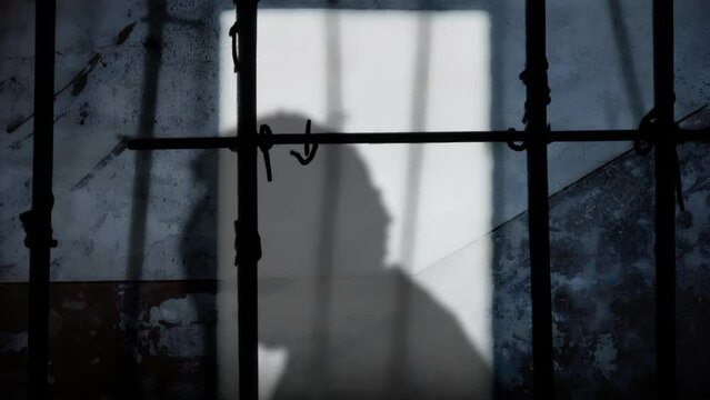 A despairing man, hands covering his face, tortured by his troubles while behind bars in a dirty prison cell, his silhouette etched on a weathered wall. Running away.
