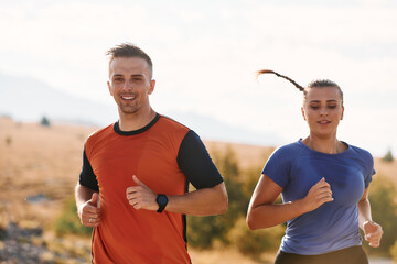 A couple dressed in sportswear runs along a scenic road during an early morning workout, enjoying...