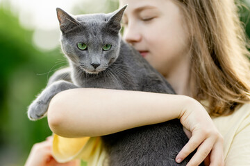 Cute teenage girl holding her pet cat on sunny summer day. Gorgeous grey Russian Blue breed cat outdoors.