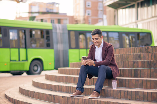 Hispanic young man sitting in a city square with his cell phone and a public bus in the background.
