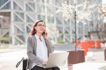 Latin young woman sitting outdoors in a city square on a sunny day with laptop and headphones,...