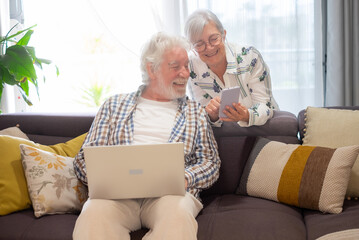 Relaxed senior white haired couple sitting on home sofa using computer and mobile phone enjoying happy retirement lifestyle in good mood