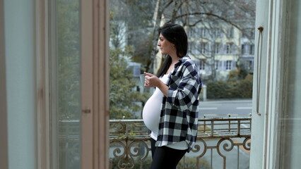 One pensive pregnant woman drinking tea by apartment balcony overlooking European city, relaxed woman enjoying view from home in third trimester pregnancy, quiet contemplation