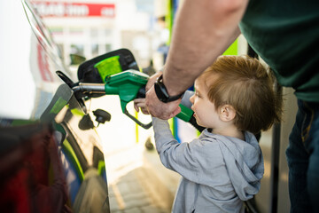 Cute little blond boy holding pump nozzle. Small kid helping father to fuel the car at a gas...
