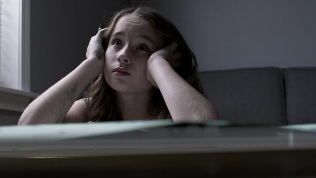 A sad young girl listens to her parents fight while she draws in a dark room