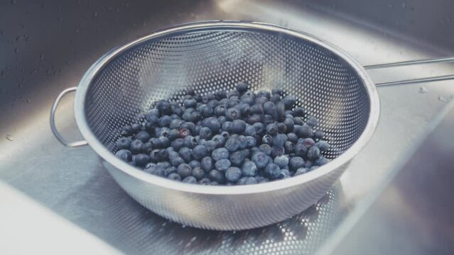 Woman fills a metal sieve with fresh blueberries and then washes blueberries with cold water in a stainless steel sink. . High quality 4k footage
