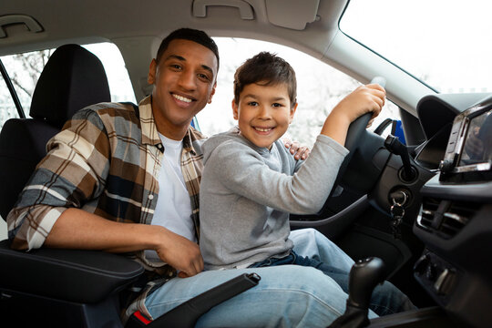Happy boy and his young father riding car together, child sitting on dad's lap in automobile, and smiling together at camera