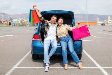 Happy spouses posing with colorful shopper bags near car outdoors, enjoy successful shopping in mall on weekend, free space, full length