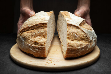 Hands holding loaf of bread cut in half with cutting board on dark worktop, close-up.