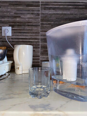 A clear glass and a water filter pitcher rest on a marble kitchen countertop, bathed in soft,...