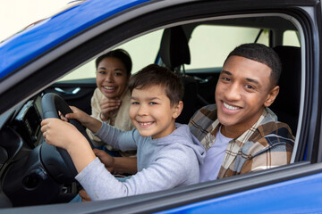 Happy preteen boy driving car, sitting with father in driver's seat smiling to camera, dad teaching...