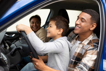 Excited preteen boy sitting on father lap, driving automobile together and smiling, enjoying their...