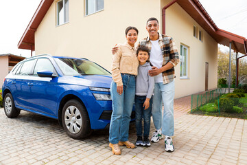 Young family of three with kid boy in front of their house and car, embracing and smiling at...