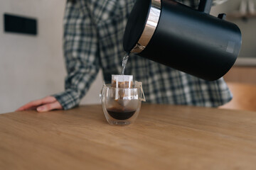 Cropped shot of unrecognizable woman pouring hot water in cup with drip coffee bag. Process of making brewed coffee at home in morning. Preparing fresh brewed drip coffee in cup with paper drip bag.