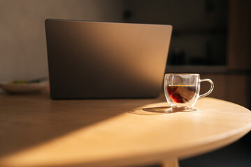 Closeup of transparent coffee cup and laptop on office desk in startup space and sunlight. Hot beverage on table in empty business workplace. Drink in glass mug on furniture and computer, no people.