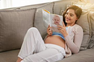 Happy and pregnant woman, reading baby book at home to prepare for motherhood on sofa. Mother,...