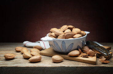 Whole almonds in ceramic bowl with cutting board and nutcracker on rustic wooden table, close-up, space for text.
