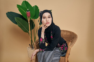 Beautiful Asian model wearing black kebaya dress with hijab, sitting on a rattan chair with bamboo...