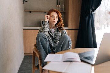 Millennial redhead businesswoman working from home office using laptop, staring at device screen looking pensive and thoughtful, search solution, prepare report or research, doing freelance job. - obrazy, fototapety, plakaty