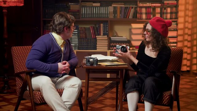 man and a woman are sitting in a vintage library in retro clothes and talking to each other about photography. An interview of two people in a pleasant, calm atmosphere.