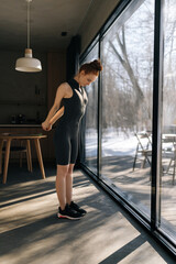 Full length vertical shot of athletic young woman in outfit preforming warming up exercise, stretching muscles before workout indoors, standing by window with snowy winter nature view on sunny day.