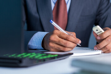 Business man wearing blue color suit and red tie writing on notepad, working on laptop