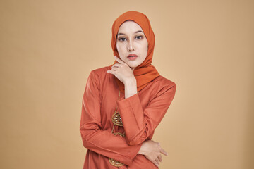 Fototapeta premium Beautiful female model wearing classic orange color kebaya with hijab, an Asian traditional dress for Muslim woman isolated over beige background. Eidul fitri festival fashion and beauty concept.