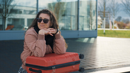 Fototapeta premium A young woman with a red suitcase is waiting for a transport, a bus outdoors. The wind blows the hair. Urban lifestyle