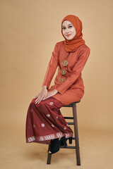 Beautiful Asian model wearing classic orange color kebaya dress with hijab, sitting on a chair isolated over beige background. Eidul fitri festival fashion and beauty concept.