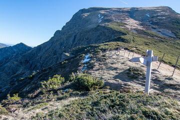 A white wooden cross stands on grassy and rocky terrain in the foreground. The background features a steep mountain slope with snow patches under a clear blue sky, likely a remote hiking trail.