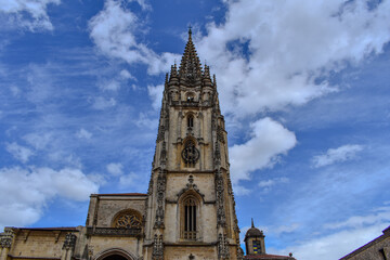 A tall, ornate church tower against a blue sky with scattered clouds. The tower features intricate Gothic architectural details, including pointed arches, decorative stonework, and a clock.