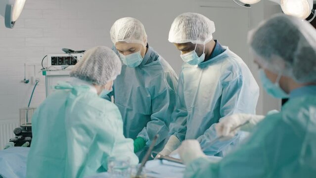 Group of mixed-races professional surgeons and nurses in uniform performing heart transplant surgery operation under bright lamps using medical instruments in operating room