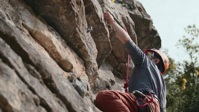 A person is climbing up a rock. They wear a helmet and hold a rope. The rock is big and the day is sunny. The climber looks up, ready to move. High quality 4k footage