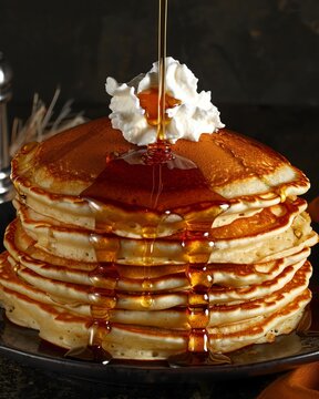 A stack of fluffy pancakes, adorned with maple syrup and whipped cream, is the centerpiece in this culinary masterpiece The golden liquid flows down like an artistic waterfall onto its soft surface, c