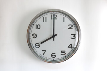 A round shape stainless steel wall clock with white face and hands set to 8 o'clock time. The clock is hanging on a wall.