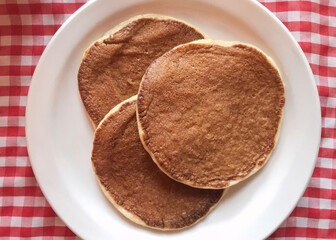 Three plain pancakes on a white plate on a red and white checkered tablecloth