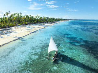 Aerial view of the sailboat on blue sea, empty white sandy beach at sunset. Summer vacation in Zanzibar. Tropical landscape with boat, ocean with clear water, green palms, sky. Top drone view. Exotic - obrazy, fototapety, plakaty
