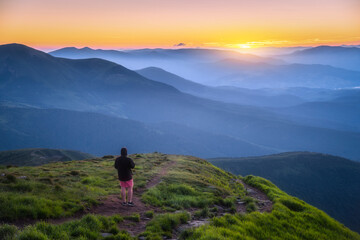 Man on the top of mountain Goverla, Ukraine. Hill with green grass and beautiful mountain valley at sunset in summer. Landscape with sporty man, hils in fog, orange sky. Hiking. Spring in Europe - obrazy, fototapety, plakaty
