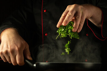 The cook adds fragrant fresh parsley by hand to a hot, steamy frying pan. Low key concept of...
