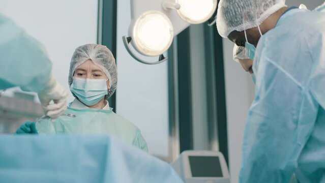 Team of professional surgeons performs surgery on patient under anesthesia in operating room. A nurse hands a medical instrument to a doctor