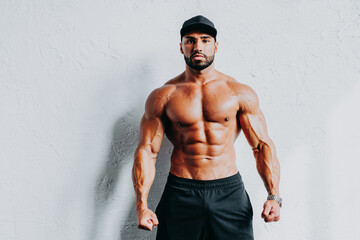 Young strong man bodybuilder in cap on white wall background