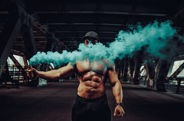 Young strong man bodybuilder standing with dense blue smoke on urban industrial background - obrazy, fototapety, plakaty