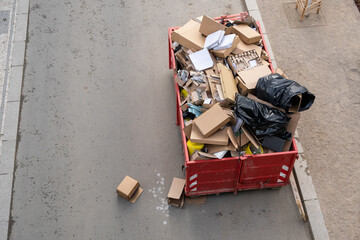 A waste container overflowed A steel industrial with rubbish on the street, Various types including empty carton boxes, plastic, and construction debris,urban waste management. construction site.