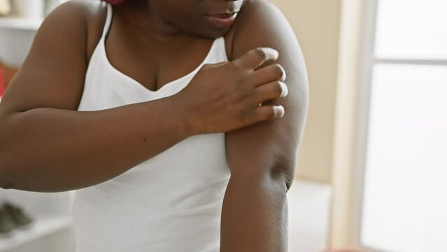 Close-up of a contemplative black woman with braids in a white tank top indoors touching her shoulder, evoking themes of beauty and self-care.