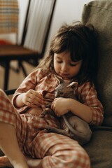 little cute girl 3 years old wearing pajamas hugging her canadian sphynx kitten sitting on a chair at home