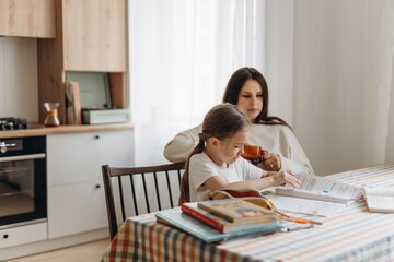 a young babysitter supervises the completion of school homework according to the lessons of the girl she looks after at her parents' home in the kitchen - obrazy, fototapety, plakaty