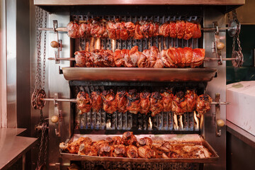 An industrial rotisserie grill oven with multiple skewers of golden-brown rotisserie pork cooking evenly over a heat source.