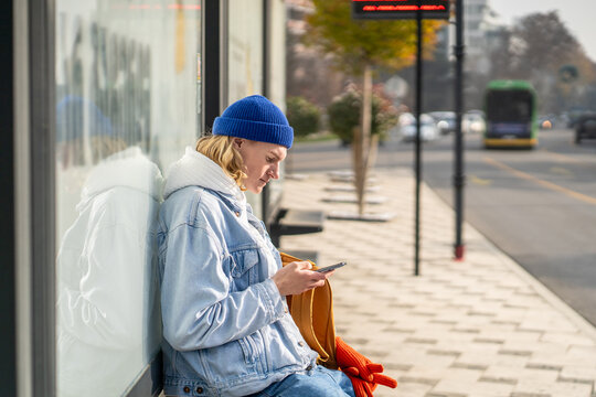 Stylish hipster, generation Z representative, sitting at bus stop, waiting for transport, messaging in smartphone, surfing in internet, scrolling web pages, photos in social networks, reading news